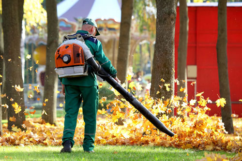 A person wearing green work clothes uses a leaf blower to clear fallen leaves in a park with trees. A red structure and carousel can be seen in the background.