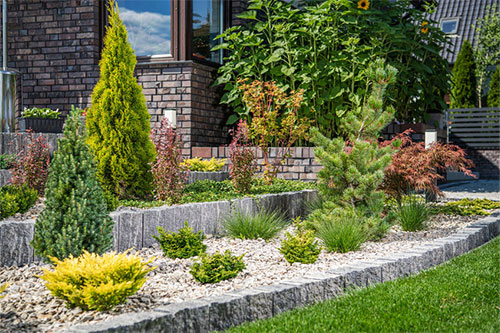 A landscaped garden features various plants and shrubs, bordered by stone retaining walls and gravel, in front of a brick house under a blue sky.