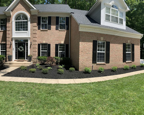 A two-story brick house with white-framed windows, black shutters, and a well-maintained front yard featuring mulched flower beds and a green lawn.