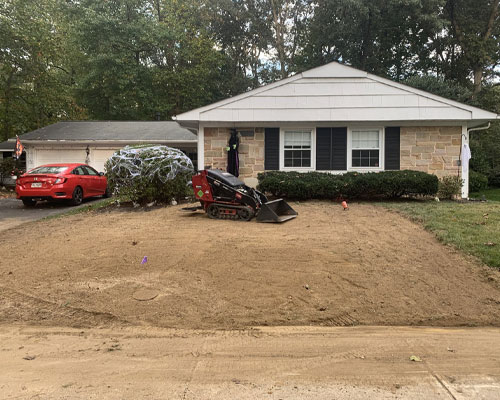 A small front yard of a suburban house with a red car in the driveway. The yard is under construction with dirt and a mini excavator. A tree covered in protective netting is visible near the house.